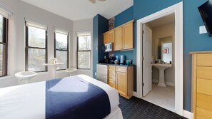 Habitación de hotel con cama, una mesita con un florero, un área de cocina con armarios y electrodomésticos, y un baño visible a través de una puerta abierta.