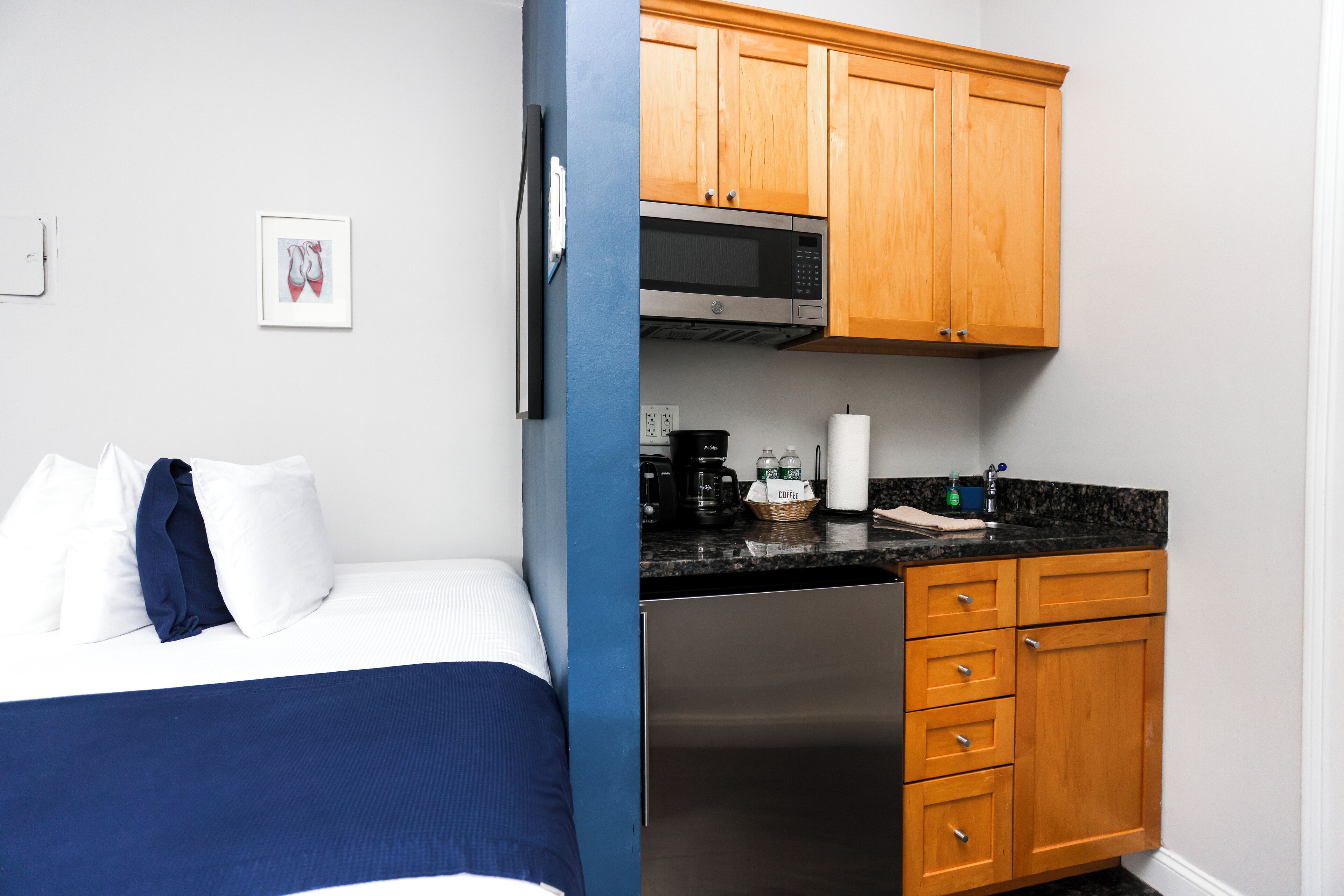 A bedroom with a bed, a blue blanket, and a white pillow. Adjacent to the bedroom is a kitchenette with wooden cabinets, a microwave, and a coffee maker.