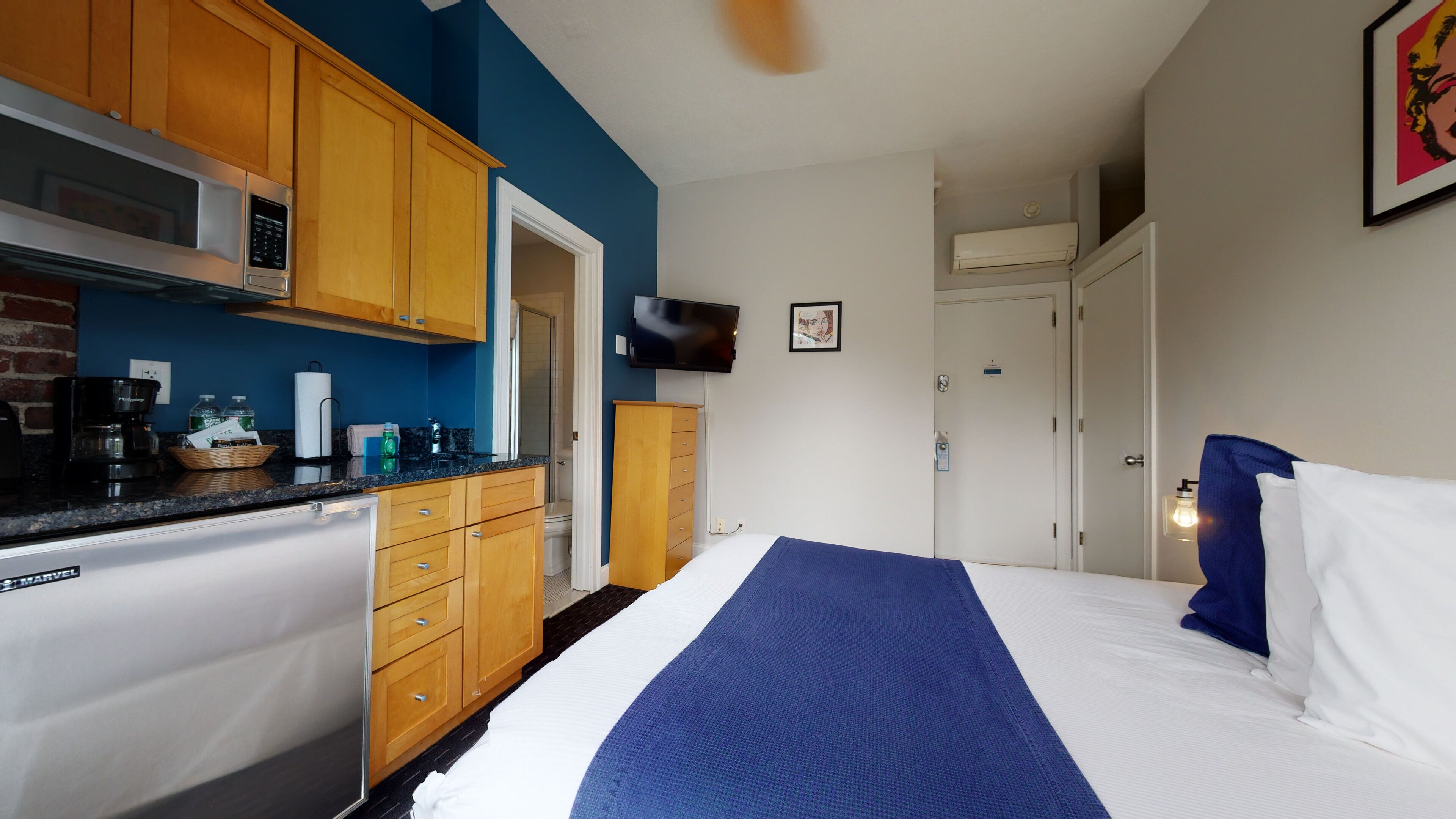 A compact hotel room with a kitchenette, a bed with a blue blanket, a flat-screen TV, and a door leading to a bathroom.
