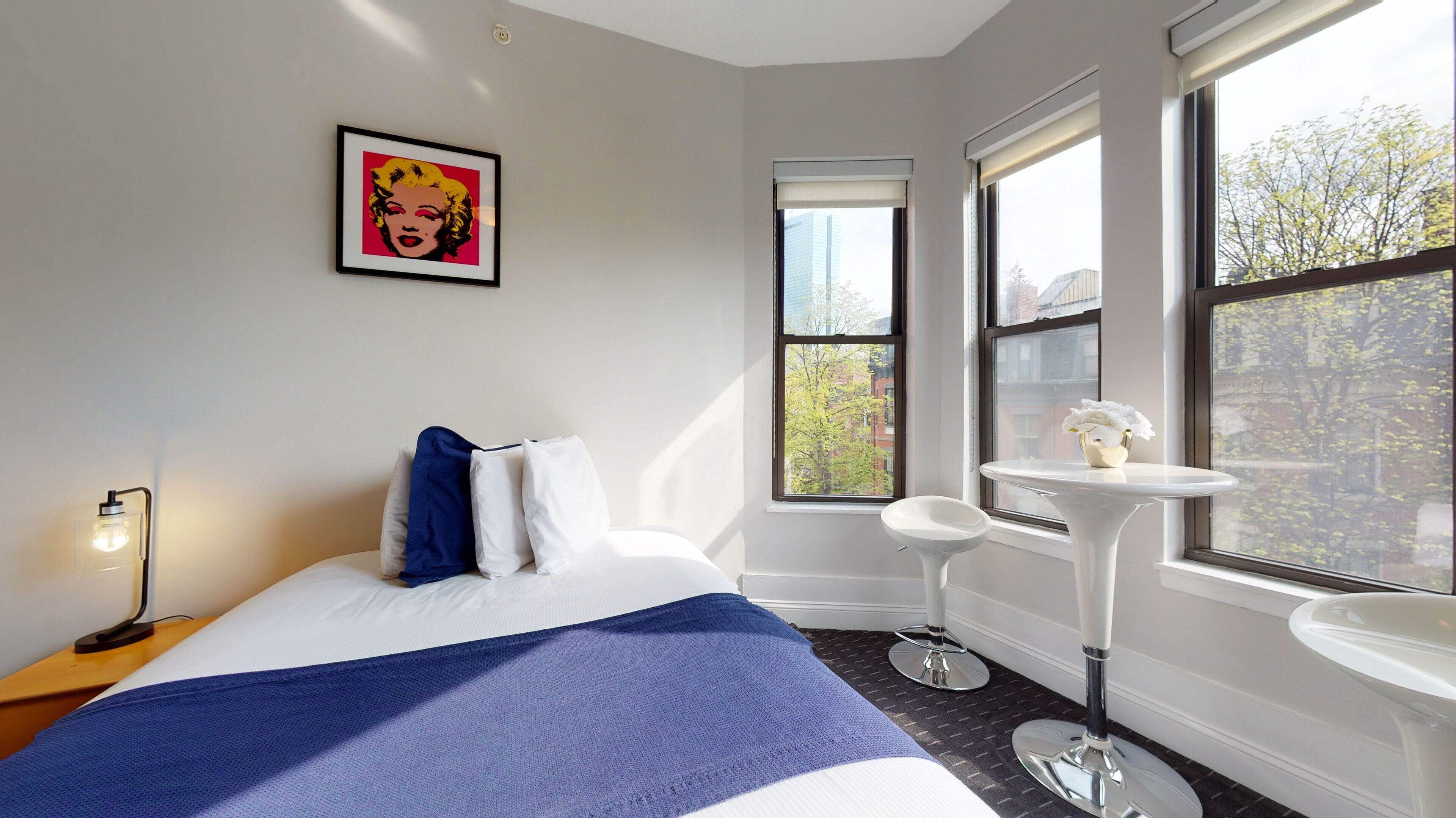 A hotel room with a bed, a bedside table with a lamp, a small white table with a vase, and large windows with a view of buildings and trees.