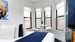 A hotel room with a large bed, a small table, two white chairs, and a window with a view of buildings. - Maverick Suites at 400 Columbus Ave (Boston)
