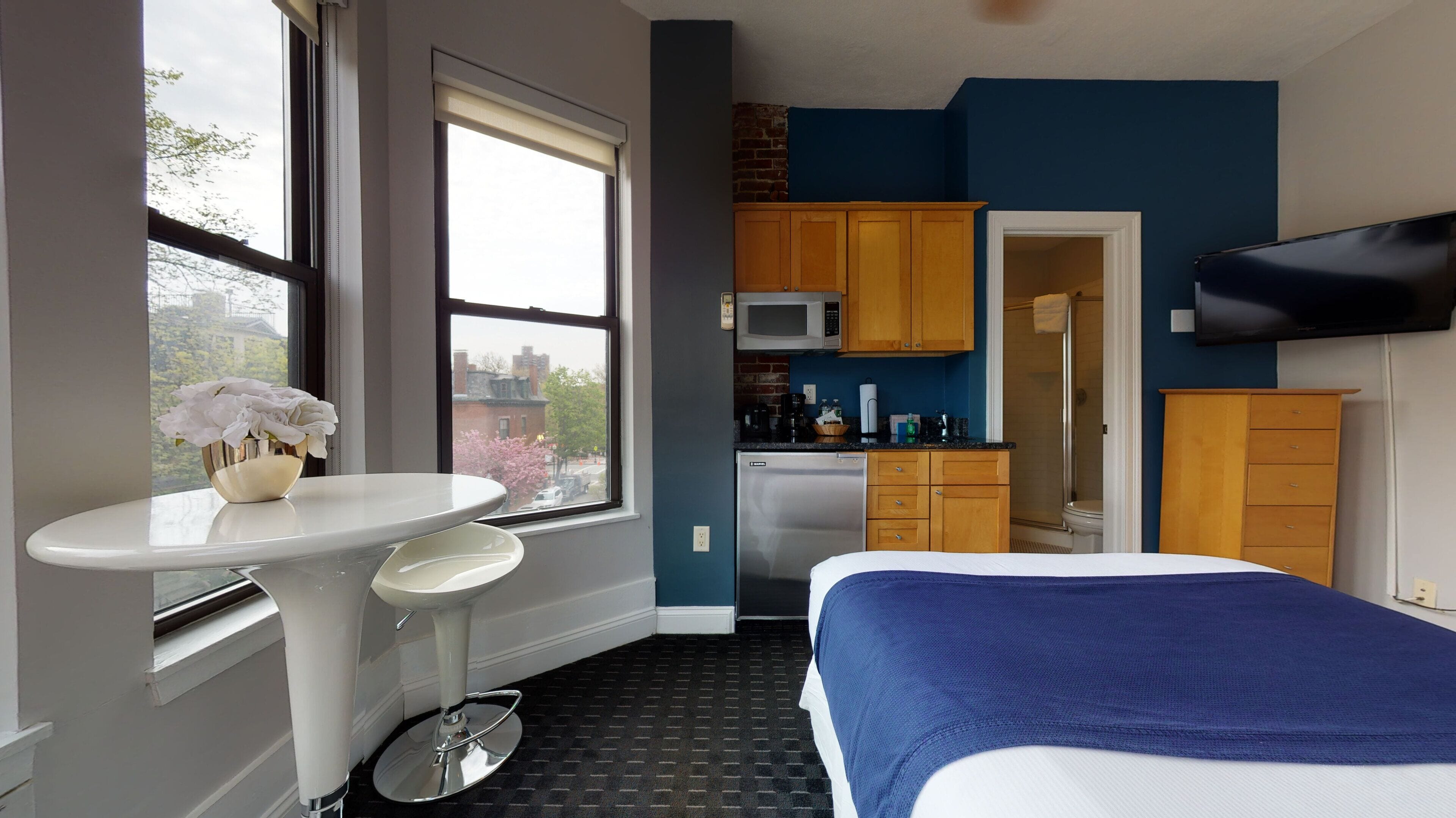 Ein kleines Hotelzimmer mit Bett, Esstisch mit Blumenvase, Küchenzeile und großem Fenster mit Blick auf die Stadt.