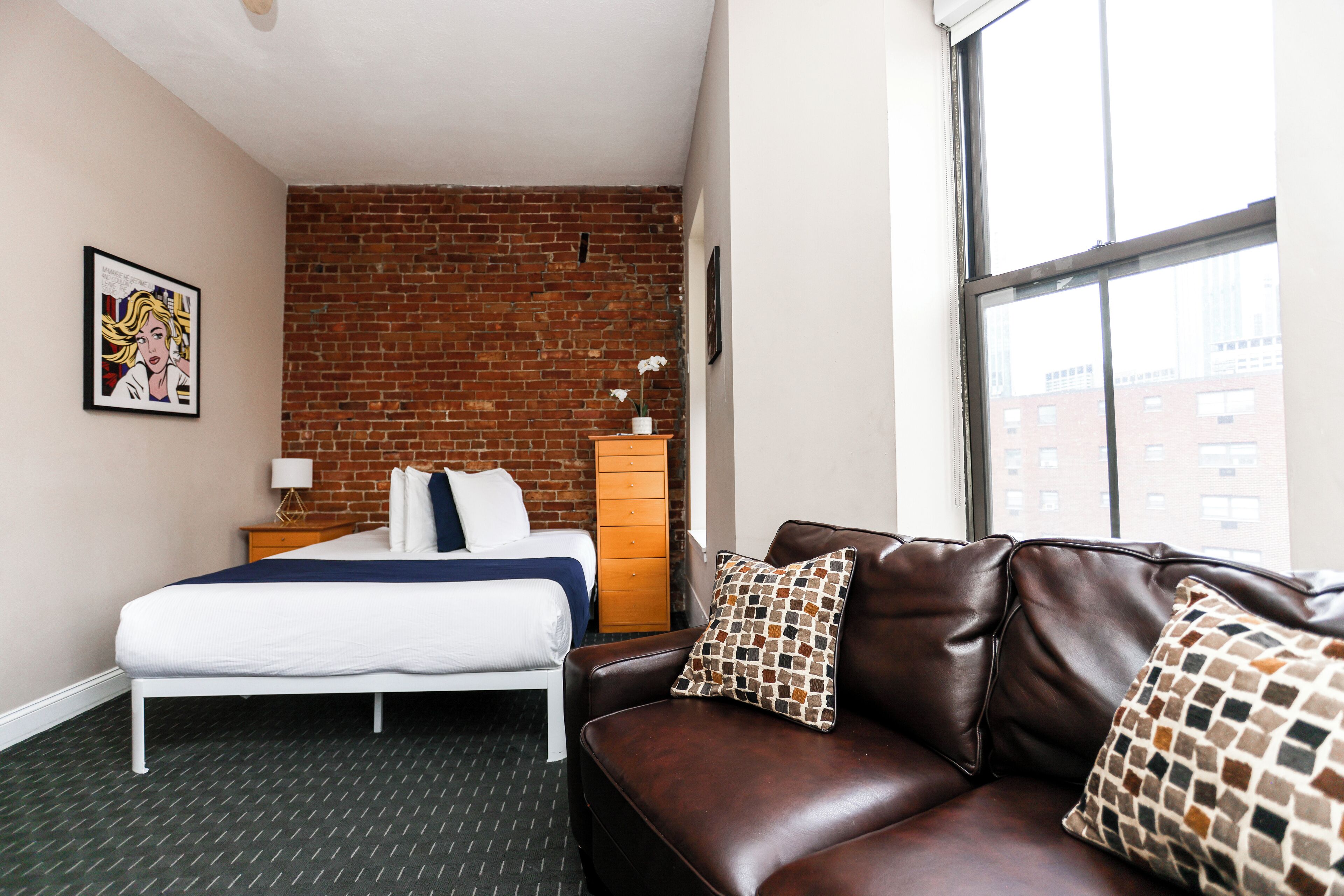 Habitación de hotel con pared de ladrillo, cama con ropa blanca, sofá de cuero marrón con cojines estampados y ventana con vista a edificios.