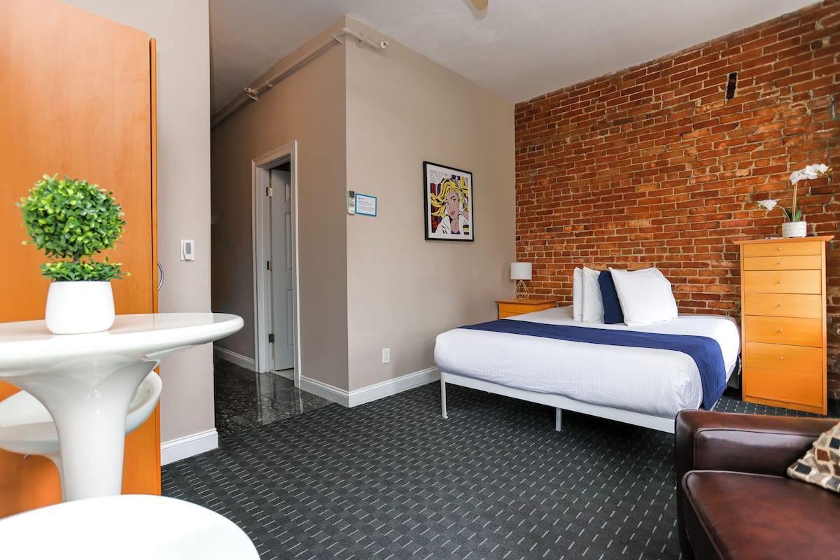 Habitación de hotel con cama, cómoda, una mesita con una planta, un cuadro enmarcado en la pared y pared de ladrillo.