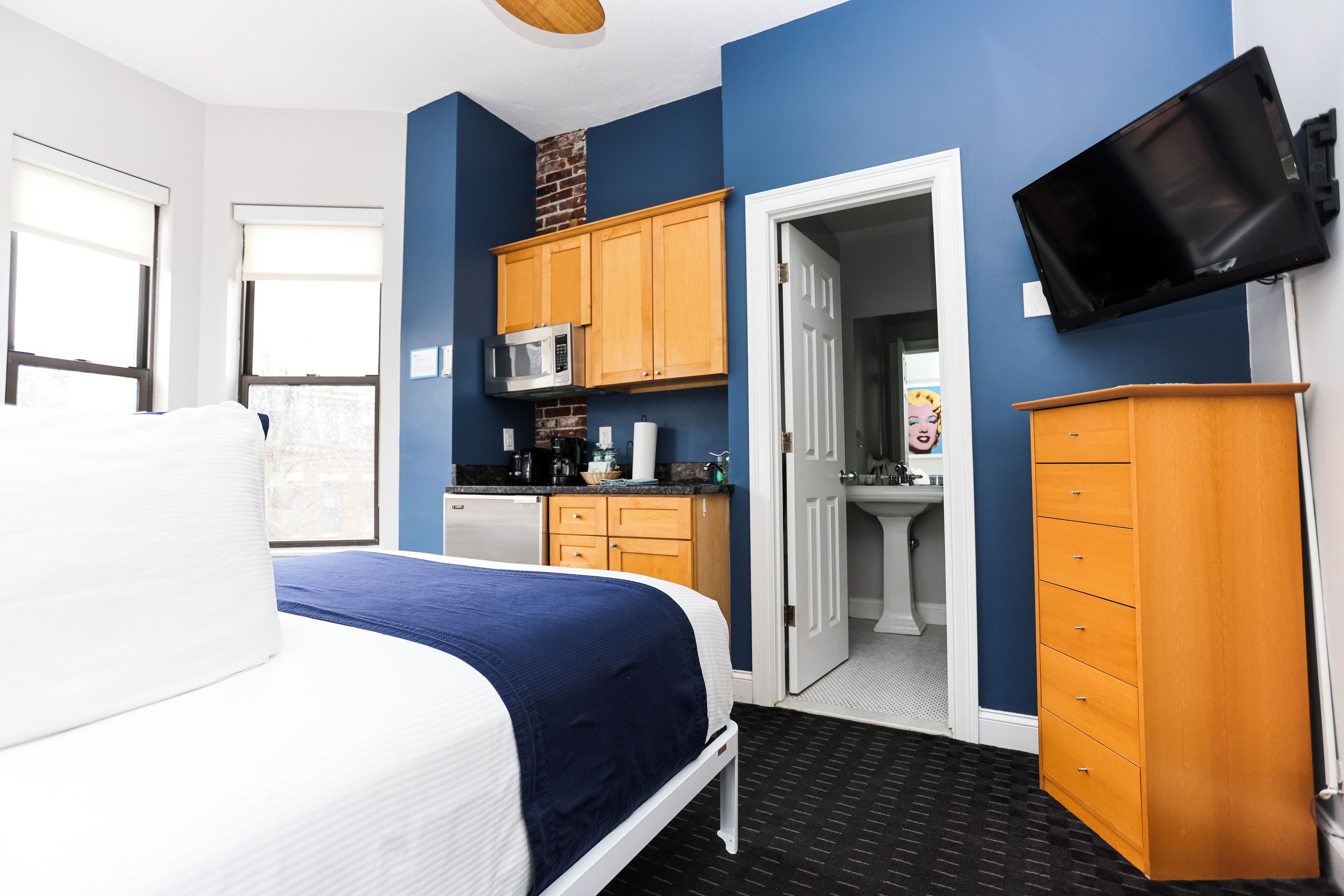 A bedroom with a bed, a television mounted on the wall, and a kitchenette area with cabinets and a microwave.