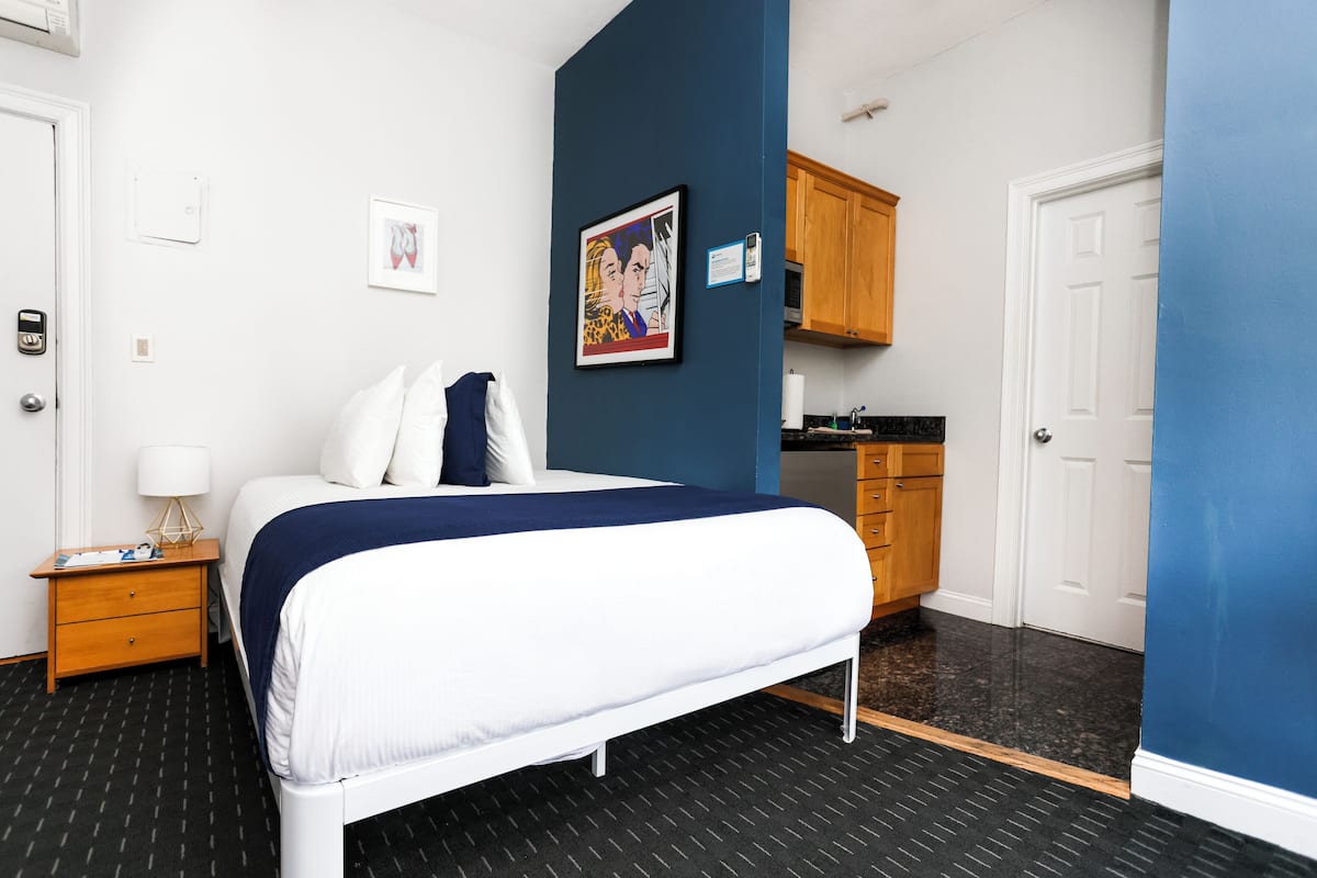 Habitación de hotel con cama, mesita de noche, un cuadro enmarcado, una puerta y una pequeña cocina.