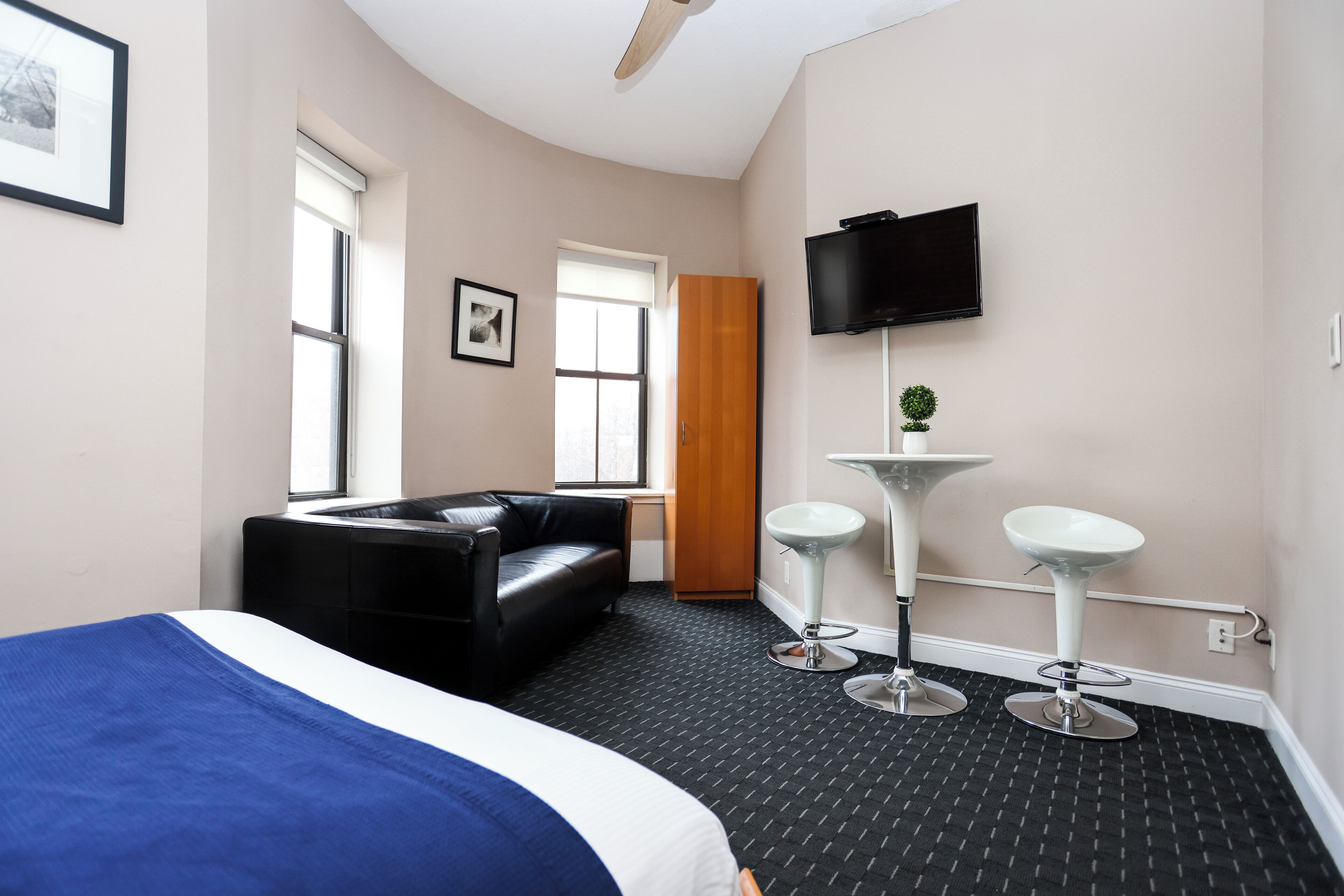 Une chambre d’hôtel avec un lit, un canapé en cuir noir, une télévision, deux tabourets de bar blancs, une petite table ronde et une fenêtre.