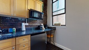 A kitchen with wooden cabinets, a black microwave, a coffee maker, and a window with a view of a brick building. - Maverick Suites at 400 Columbus Ave (Boston)