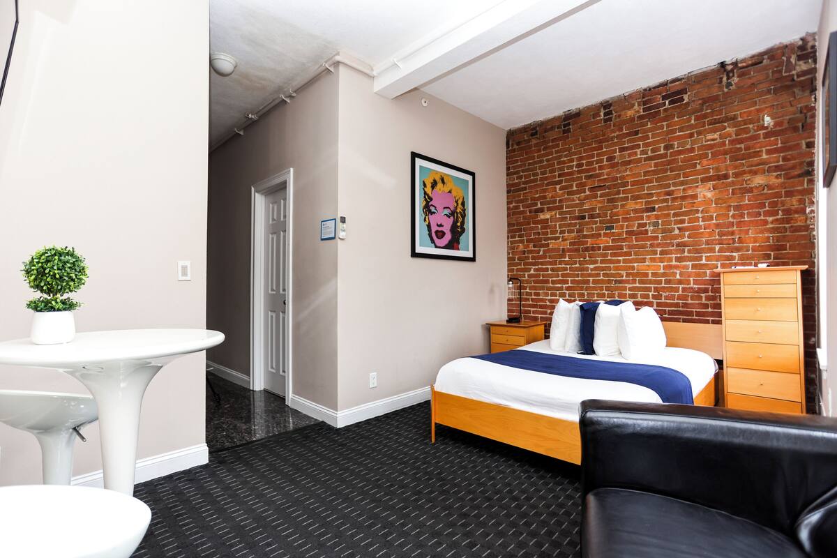 Habitación de hotel con pared de ladrillo, cama con ropa de cama blanca y azul, cómoda de madera, sillón de cuero negro y una pequeña mesa blanca con planta en maceta.