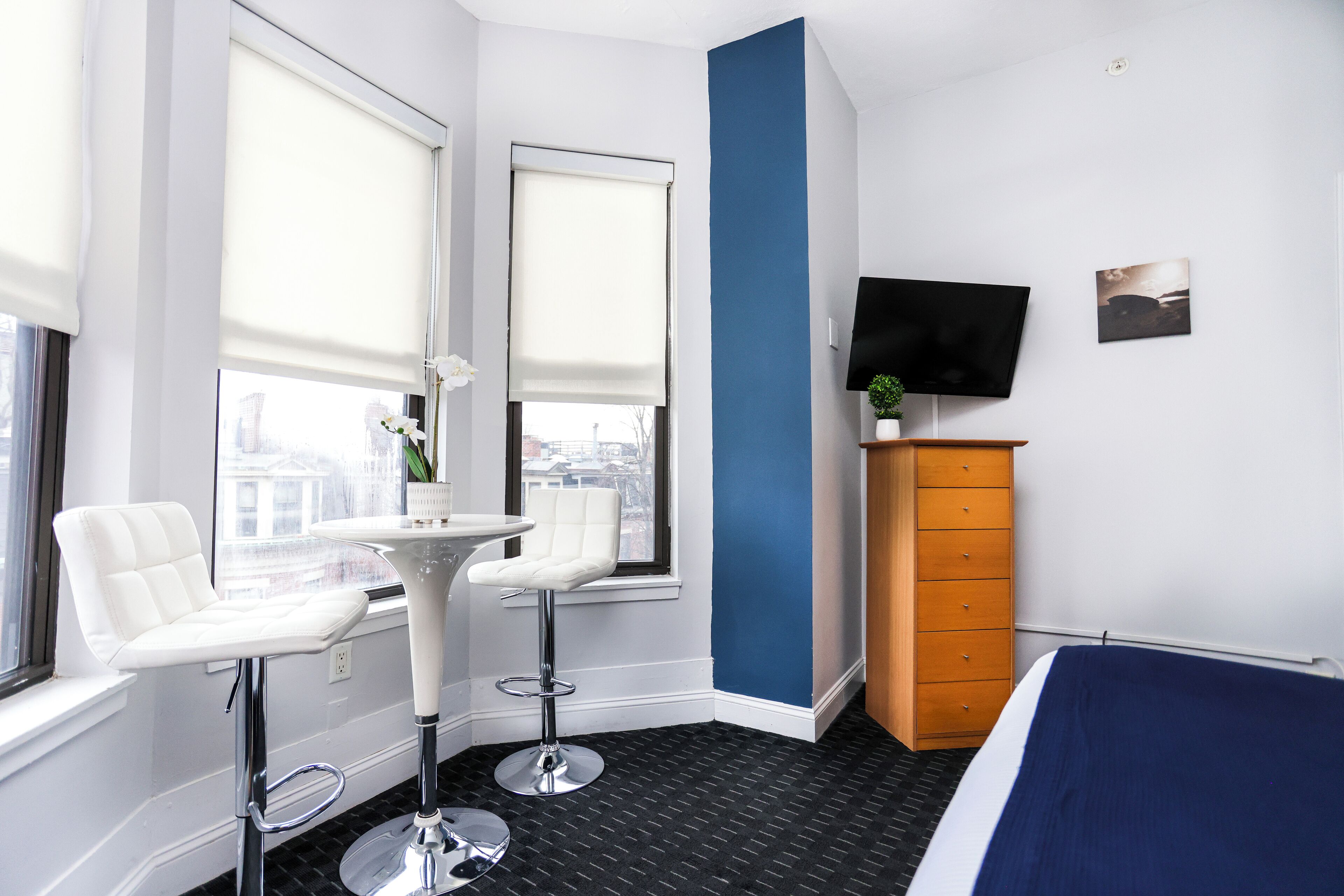 A hotel room with a bed, two white bar stools, a small table, a TV, and a dresser.