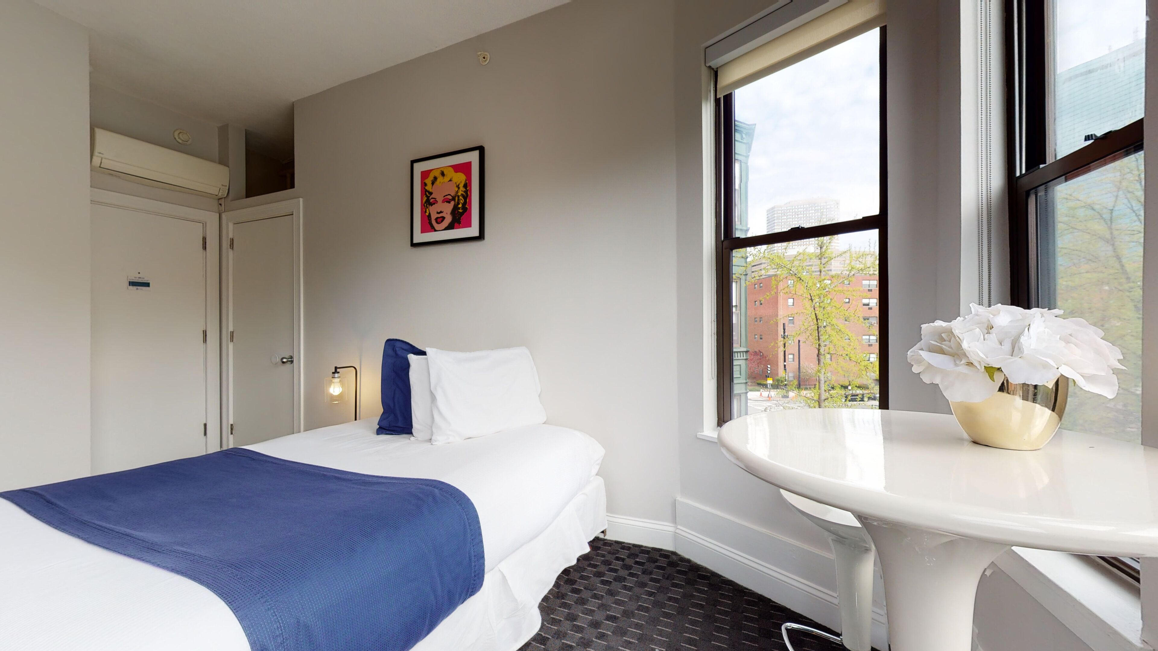 A hotel room with a bed, a bedside table with a vase of flowers, a window with a view of buildings, and a door.