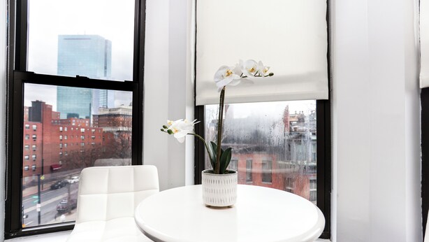 A white table with a vase of white orchids, a white chair, and a view of a cityscape through a window with a black frame. - Maverick Suites at 400 Columbus Ave (Boston)
