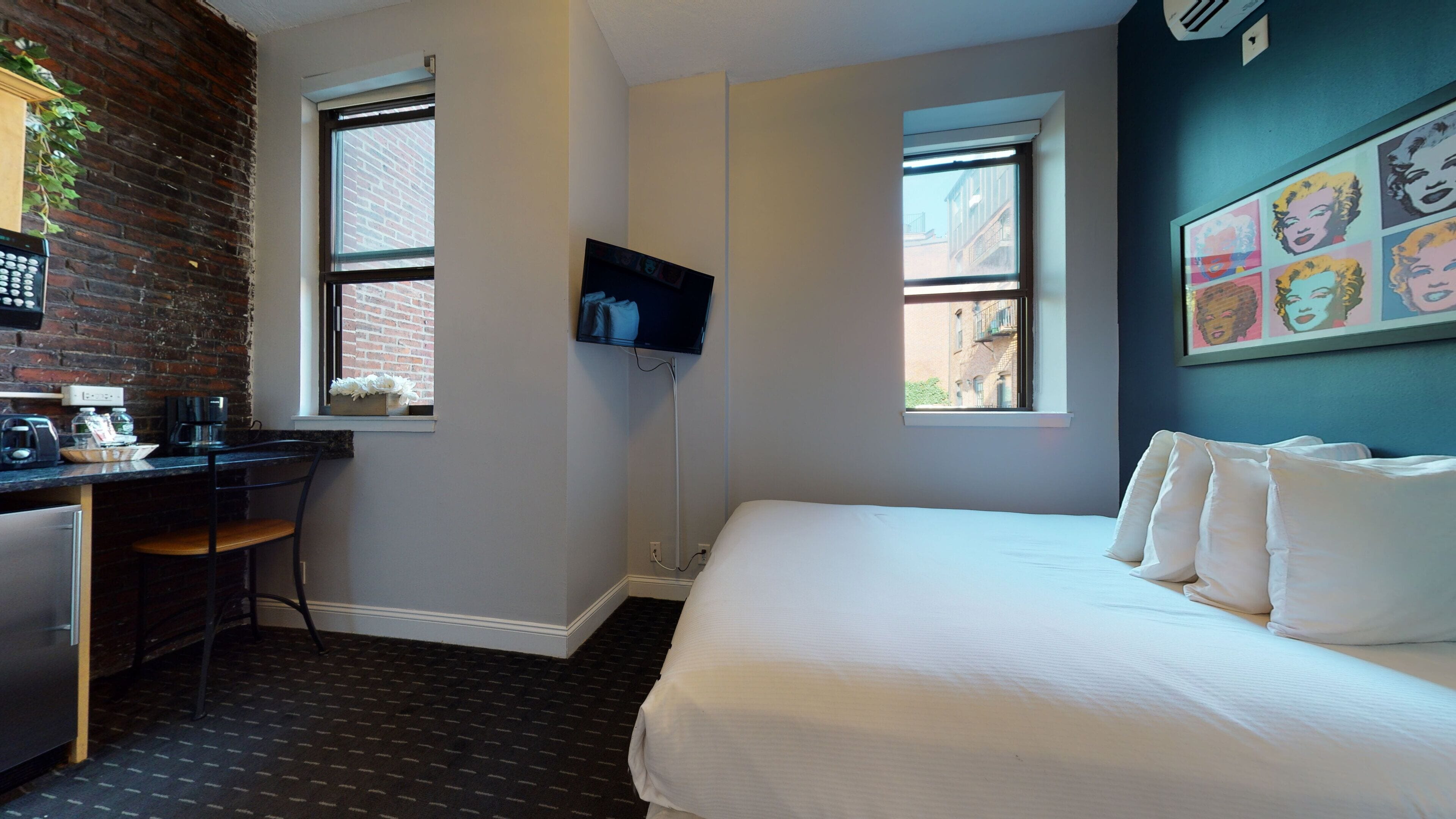 A hotel room with a bed, a desk with a chair, a TV mounted on the wall, and a window with a view of buildings.
