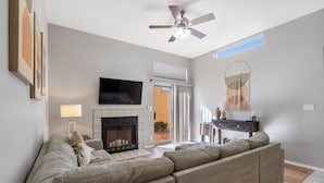 A living room with a sectional sofa, a fireplace, a ceiling fan, and a flat-screen TV mounted on the wall.