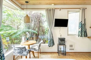 Um quarto com uma mesa de madeira redonda, duas cadeiras, uma televisão na parede e uma janela com cortinas.