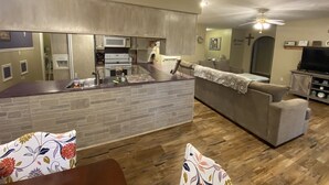 Una cuina moderna amb forns i microones integrats, una zona de menjador amb taula de fusta i coixins florals, i un saló amb sofà i televisió.