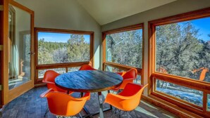 A room with a round table and orange chairs, large windows, and a view of trees.