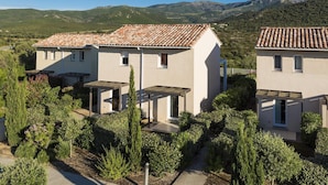A modern two-story house with a tiled roof, surrounded by lush greenery and mountains in the background.
