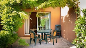 Eine Terrasse im Freien mit Tisch und Stühlen, umgeben von Grünpflanzen und einer Glastür, die zu einem Zimmer führt.