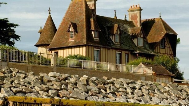 A large, historic building with multiple steeply pitched roofs and a stone wall in the foreground. - Escapade à Grandcamp av/Piscine, Sauna et Wifi (Grandcamp-Maisy)