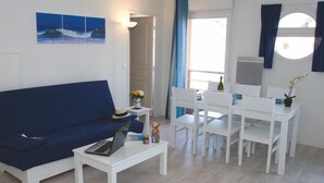 A living room with a blue sofa, a white coffee table, and a dining area with white chairs and a table. - Escapade à Grandcamp av/Piscine, Sauna et Wifi (Grandcamp-Maisy)