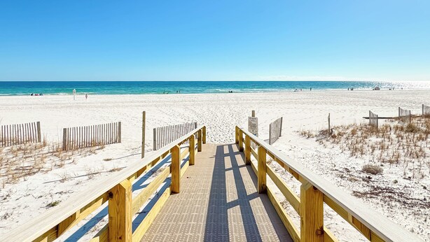 Ein hölzerner Steg führt zu einem Sandstrand, der von einem klaren, blauen Himmel gesäumt wird.