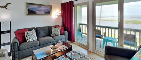 Una sala de estar con un sofá gris, una mesa de centro y vista a la playa a través de grandes puertas correderas de vidrio.