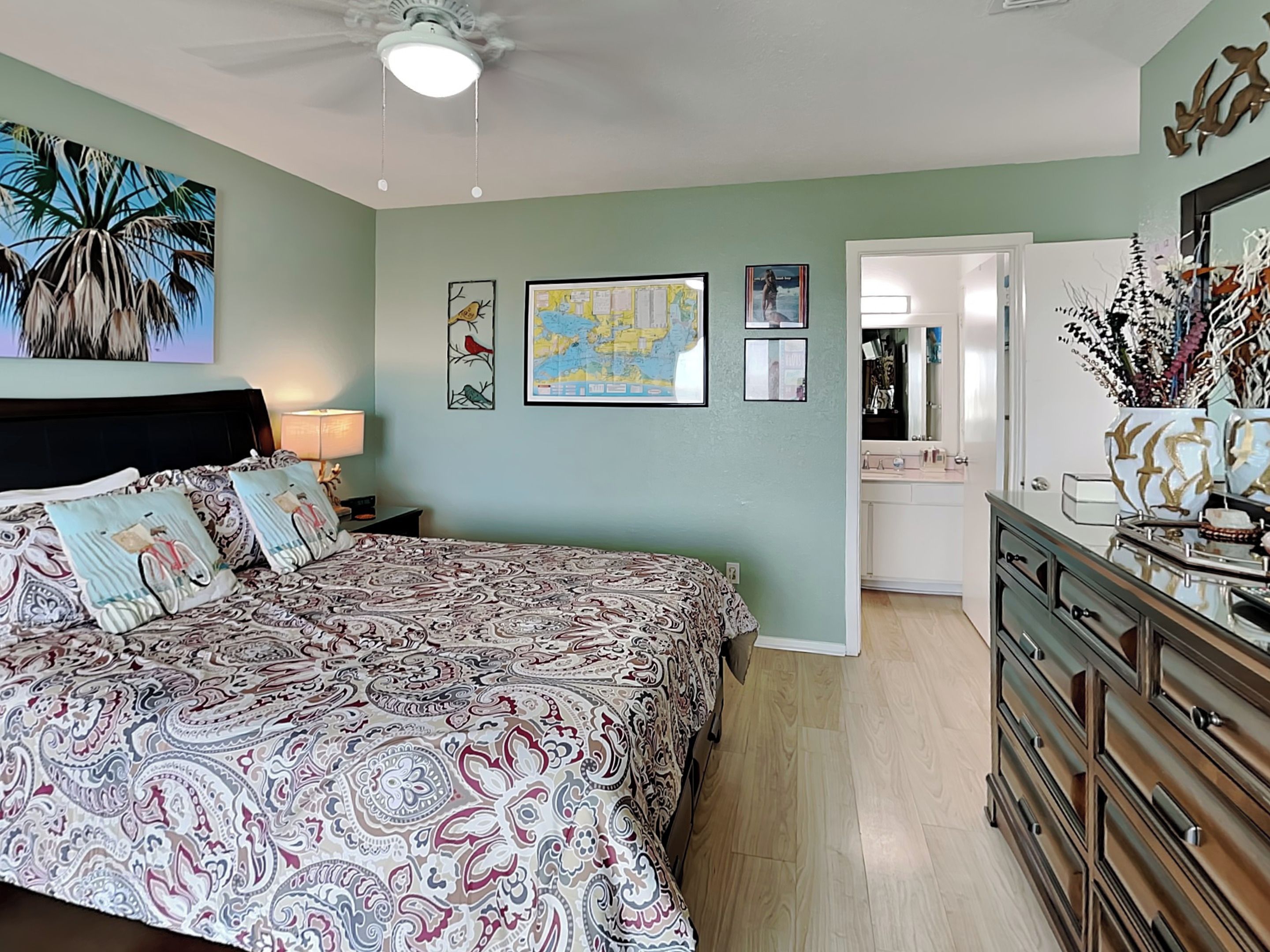Une chambre à coucher avec un grand lit, une commode, un ventilateur de plafond et une salle de bain visible à travers une porte ouverte.