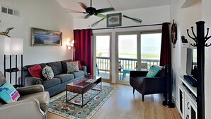 A living room with a sofa, armchair, coffee table, and a TV. There is a balcony with a view of the ocean. - Golden Gull by Miss Kitty’s (Rockport)