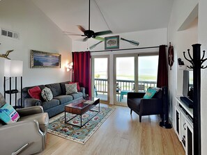Ett vardagsrum med en soffa, fåtölj, soffbord och en tv. Det finns en balkong med utsikt över havet.