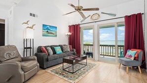 A living room with a sofa, armchair, and a coffee table. There is a ceiling fan, a painting, and a view of the outdoors through a sliding door. - Golden Gull by Miss Kitty’s (Rockport)