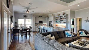 A spacious living room with a gray sectional sofa, a dining area with a wooden table and chairs, and a kitchen with white cabinets and a built-in microwave.