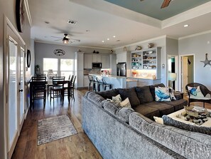 A spacious living room with a gray sectional sofa, a dining area with a wooden table and chairs, and a kitchen with white cabinets and a built-in microwave. - Tequila Sunset by Miss Kitty’s (Rockport)