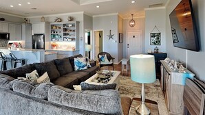 A modern living room with a grey sectional sofa, a coffee table, and a lamp. There is a kitchen in the background with white cabinets and a dining area with a table and chairs.