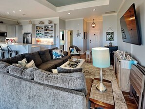 A modern living room with a grey sectional sofa, a coffee table, and a lamp. There is a kitchen in the background with white cabinets and a dining area with a table and chairs. - Tequila Sunset by Miss Kitty’s (Rockport)