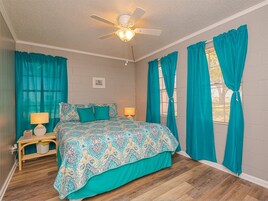 Un dormitorio con cama, cortinas color turquesa, ventilador de techo y mesita de noche con lámpara.