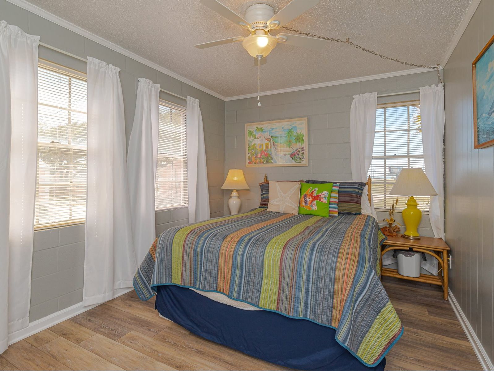 A bedroom with a bed, two bedside lamps, a ceiling fan, and a painting on the wall.