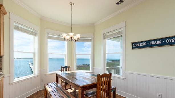 A dining room with a wooden table and chairs, large windows with ocean views, and a sign on the wall listing seafood.