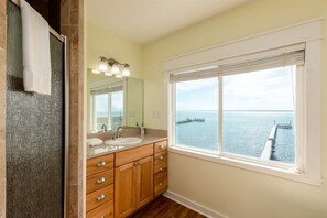 A bathroom with a double sink, wooden cabinets, a mirror, and a window with a view of the sea and a pier. - Sea Forever by Miss Kitty’s (Rockport)