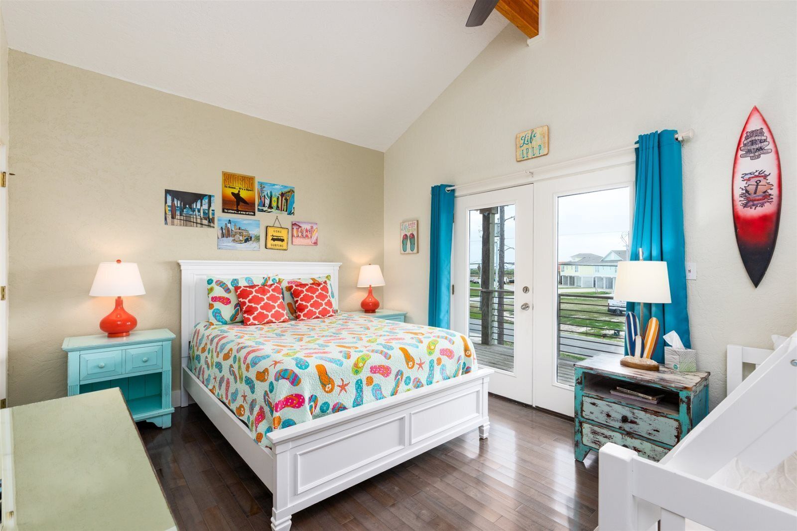 A bedroom with a bed, two nightstands, a window with curtains, and a surfboard on the wall.