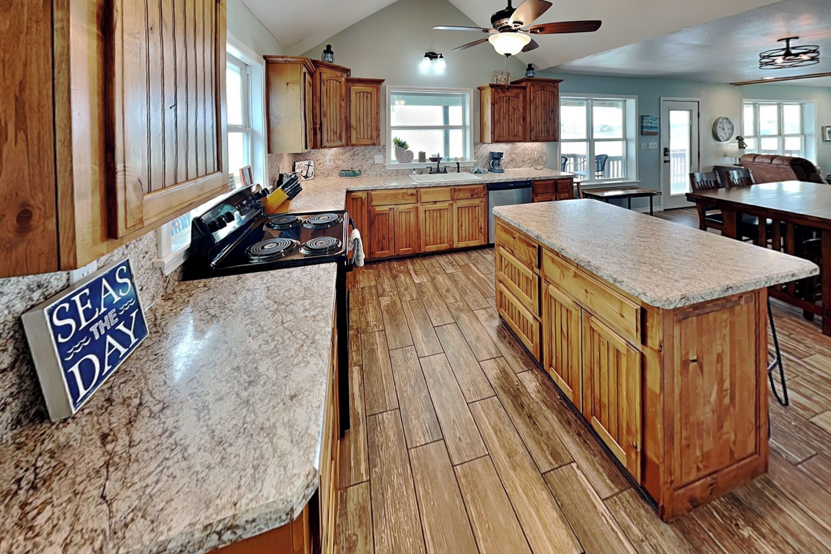 A spacious kitchen with wooden cabinets, a granite countertop, and a sign that reads 'SEAS THE DAY'.