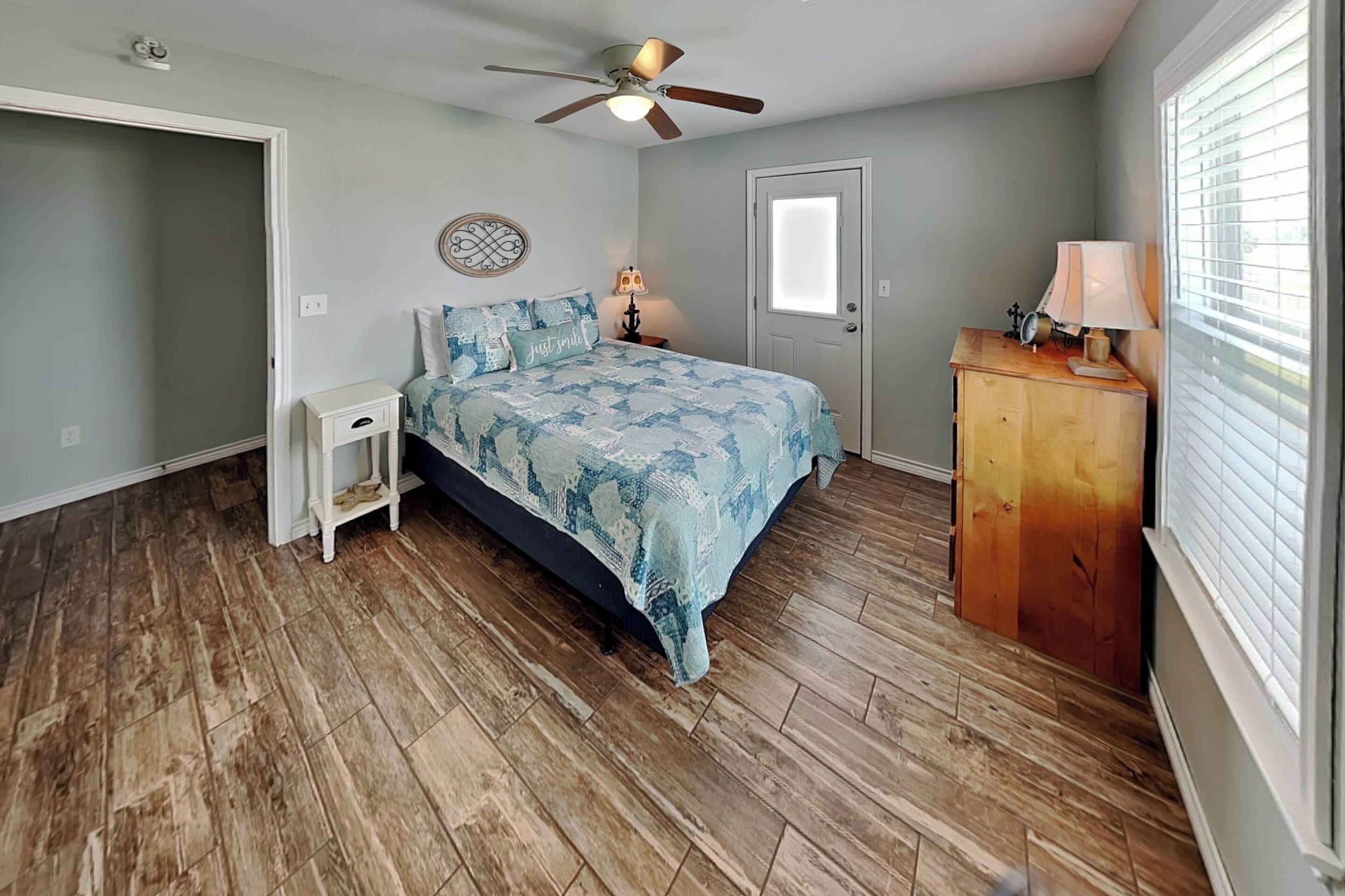 Une chambre à coucher comprenant un lit, une table de chevet, une commode, un ventilateur de plafond et une porte.