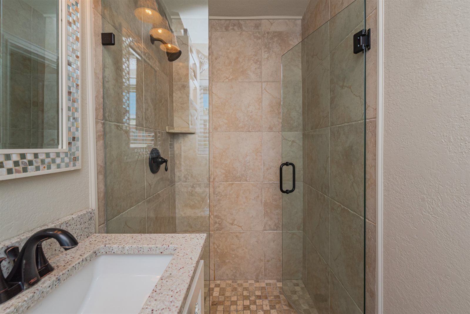 A modern bathroom with a glass shower enclosure, a granite countertop, and a mosaic tile backsplash.