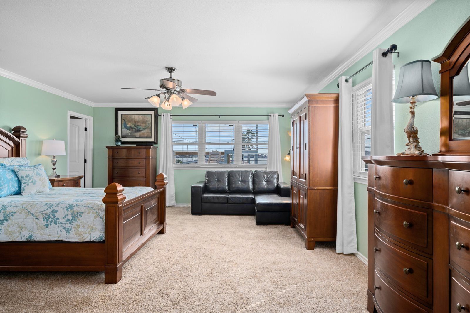 A bedroom with a bed, a dresser, a nightstand, a ceiling fan, a window with blinds, and a view of a building outside.
