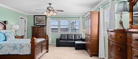 A bedroom with a bed, a dresser, a nightstand, a ceiling fan, a window with blinds, and a view of a building outside.