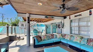 A covered outdoor seating area with a ceiling fan, a blue sofa with patterned cushions, and a checkered rug. - Texas Tiki by Miss Kitty’s (Rockport)