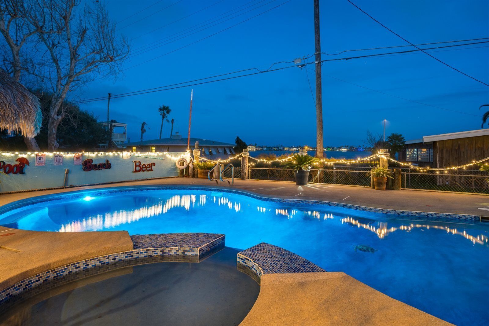 A swimming pool with a tiled edge, surrounded by a deck area with string lights and a sign that reads "Beer."
