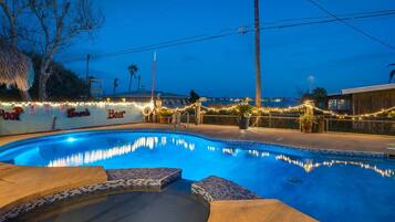 A swimming pool with a tiled edge, surrounded by a deck area with string lights and a sign that reads "Beer."