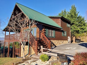 A log cabin with a green metal roof, wooden deck, and a stone wall.