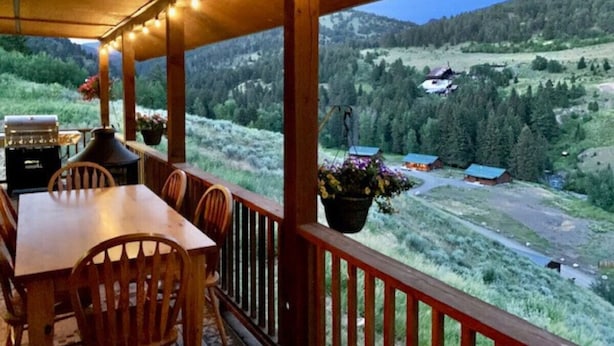 A wooden deck with a table and chairs, overlooking a mountainous landscape with a view of a small cabin and forested area.